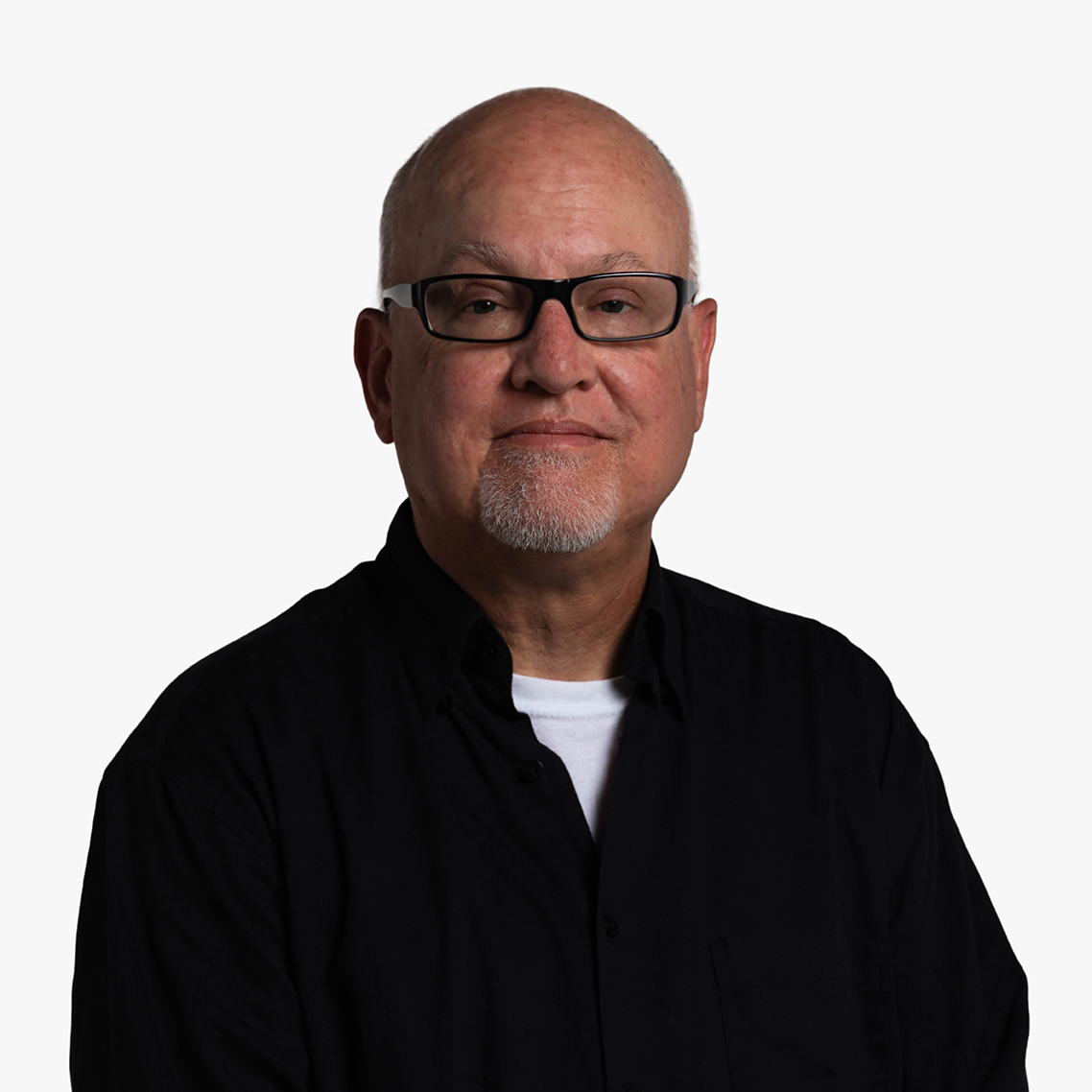 Lewis Francis, an older balding white dude with glasses and a white goatee, standing in requisite black with a white T-shirt exposed at open collar, hands clasped in front and looking directly at the camera with bemused expression.