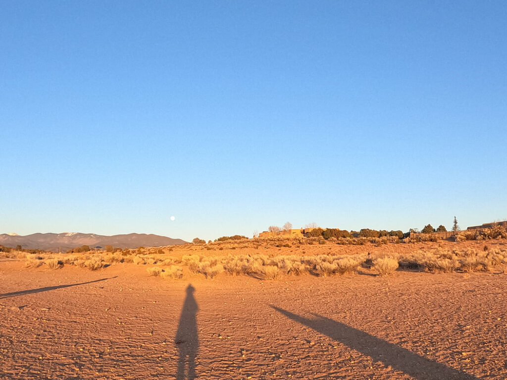 Dusty desert landscape in Santa Fe, New Mexico, with long shadows in the foreground and mountains in the distance under the bright blue sky with the moon visible low on the horizon