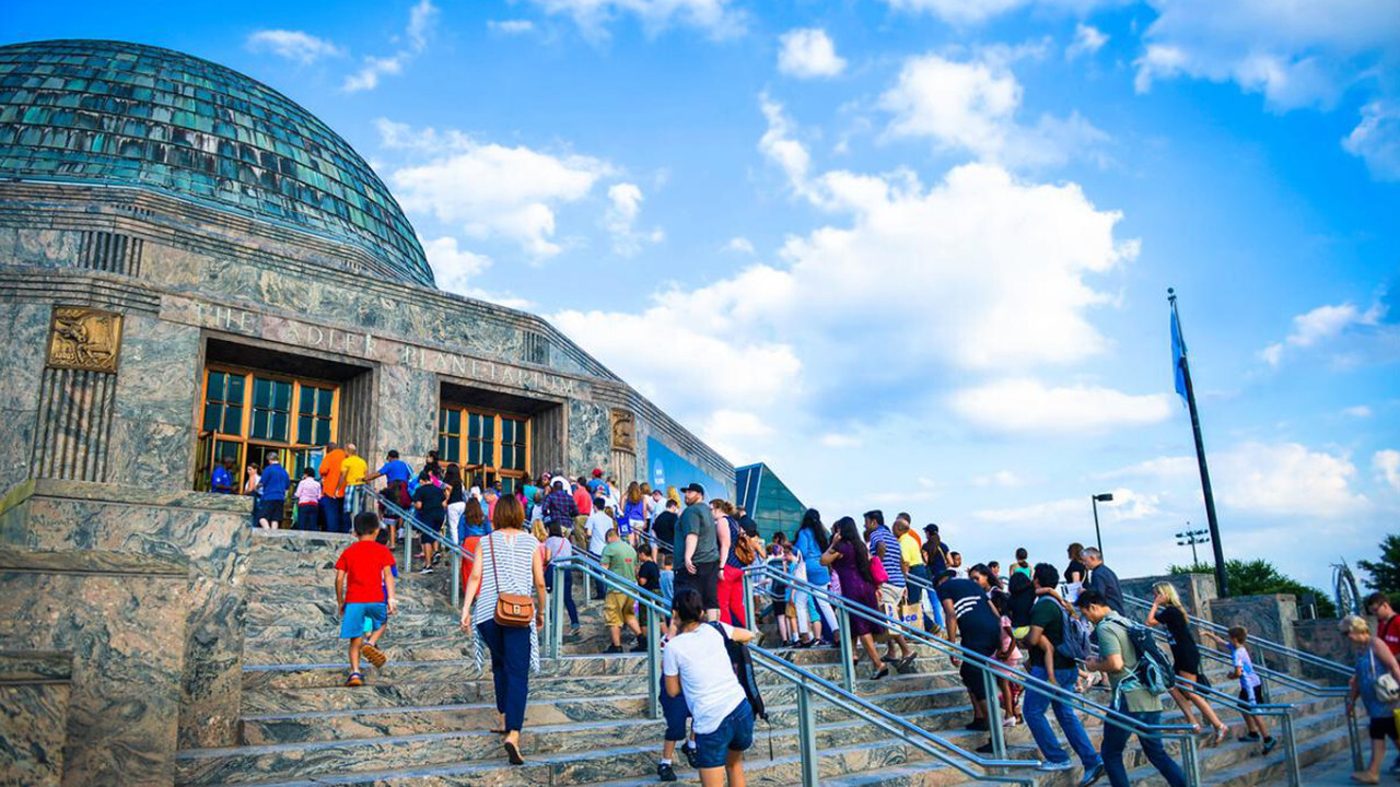 Visitors line up on the steps outside the Adler Planetarium building under a blue sky.